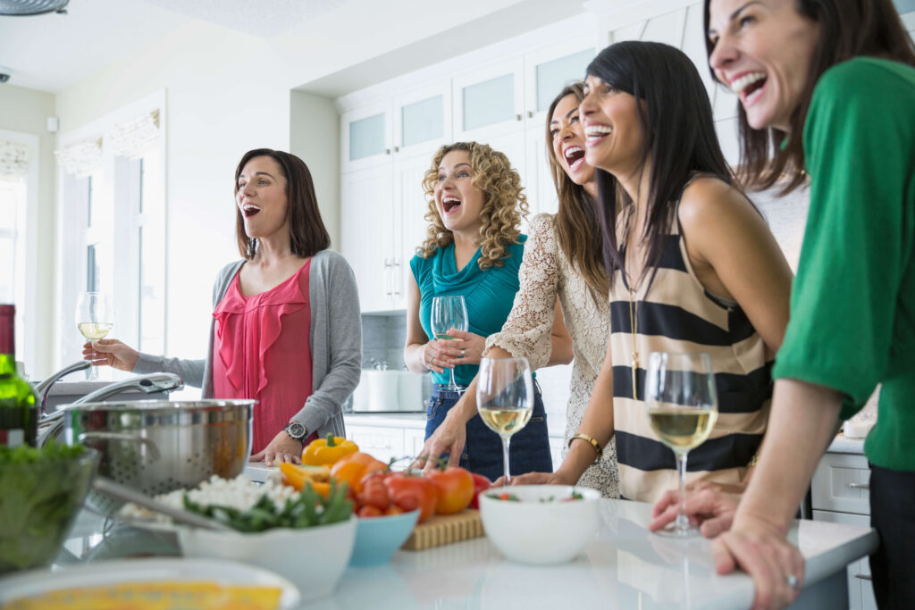 5 women drinking wine and laughing around a kitchen island